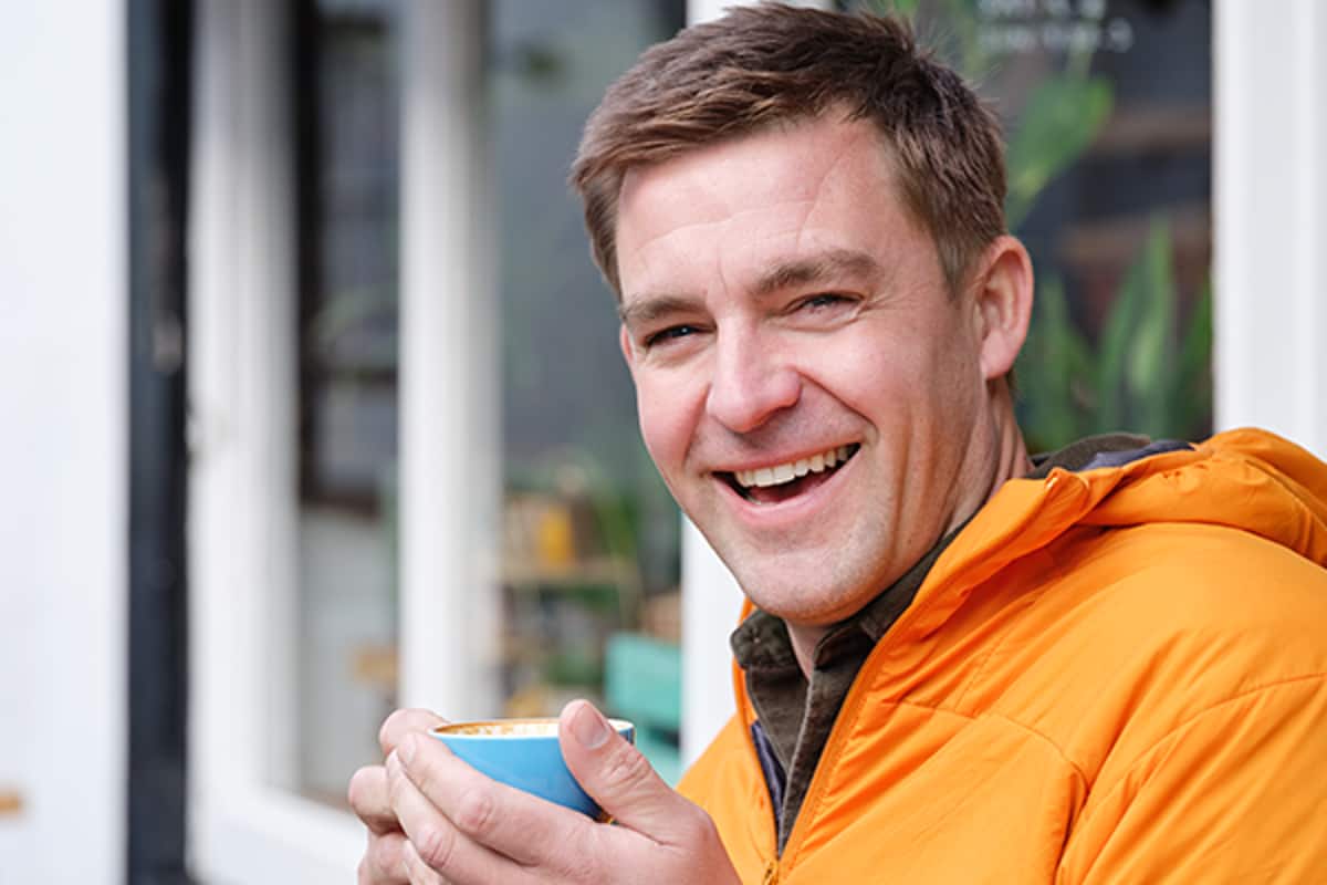 Mann mit orangener Jacke und Tasse in der Hand lacht
